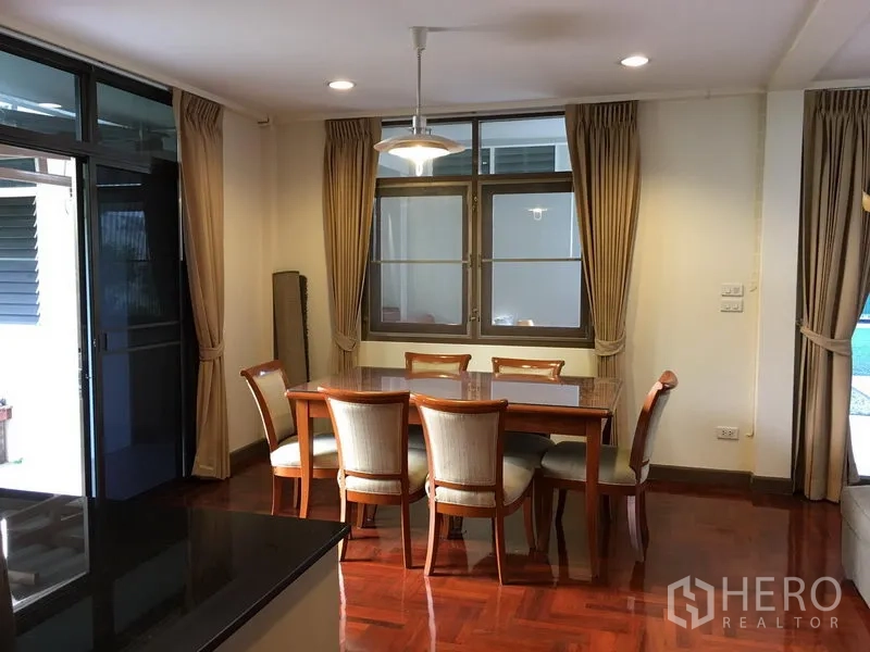 Detached House for rent in Khlong Toei, Bangkok - Dining area with wooden table and chairs, large windows and neutral curtains next to the kitchen.