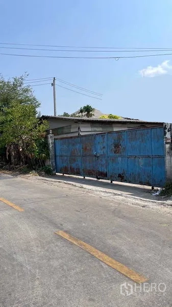 คลังสินค้า ให้เช่าในNong Khaem, Bangkok - มุมมองหน้าทางเข้าโกดังพร้อมสายไฟเหนือศีรษะและต้นไม้ริมถนนในย่านหนองแขม