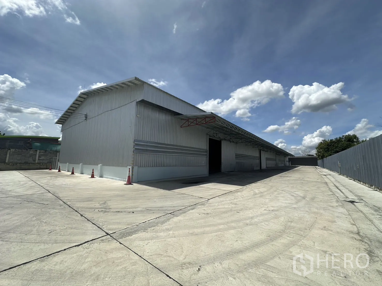 Warehouse for rent in Mueang Pathum Thani, Pathum Thani - Side view of the warehouse within a fenced compound, showing extended concrete driveway and safety cones.