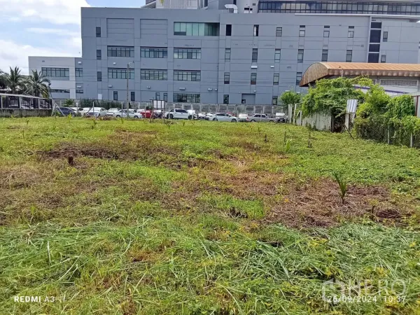 Land for rent in Khan Na Yao, Bangkok - Cleared plot with young plants beside a large modern industrial or office building and parking area.