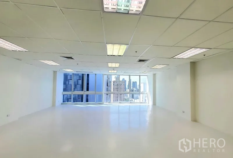 Office Space for rent in Khlong Toei, Bangkok - Spacious 110 sqm office space with grid ceiling, fluorescent lighting, and floor‑to‑ceiling windows overlooking the city.