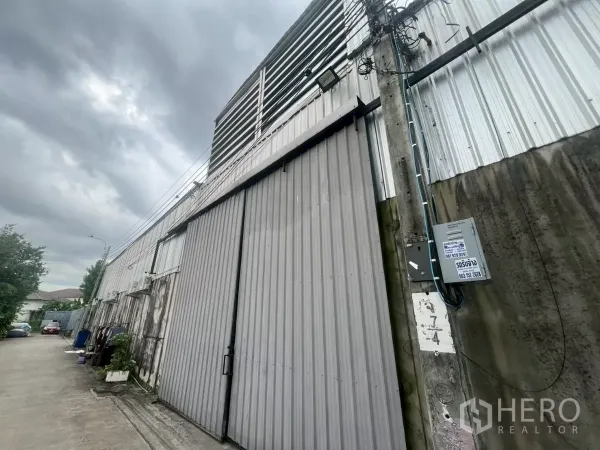 货栈 出租 - 临街金属外墙的仓库立面，超宽推拉门与电线杆，天空多云。