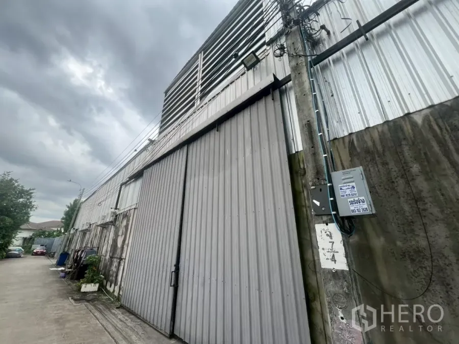 货栈 出租 - 临街金属外墙的仓库立面，超宽推拉门与电线杆，天空多云。