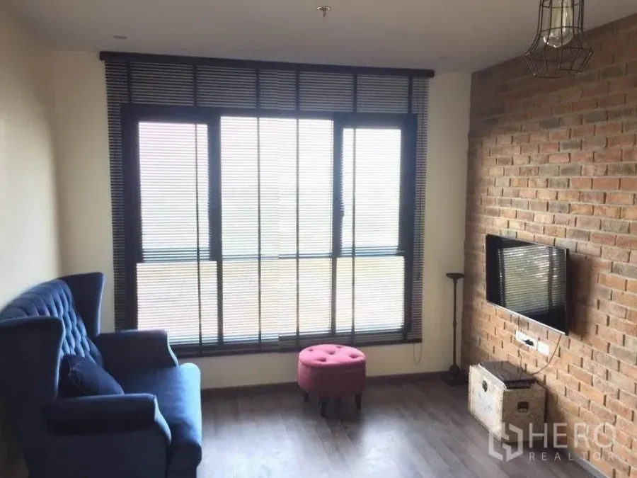 Condominium for sale in Watthana, Bangkok - Bright living room with large windows and blinds, exposed brick TV wall, blue armchair and dark wood flooring.