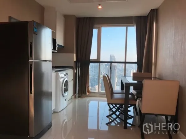 Condominium for rent or sale in Huai Khwang, Bangkok - Open-plan kitchen with fridge, washer and a dining set facing a large city-view window.