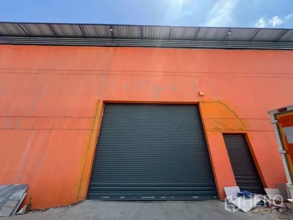 货栈 出租或出售在Lat Krabang, Bangkok - 橙色仓库立面，配大型卷闸门与小型侧门，金属屋顶下。