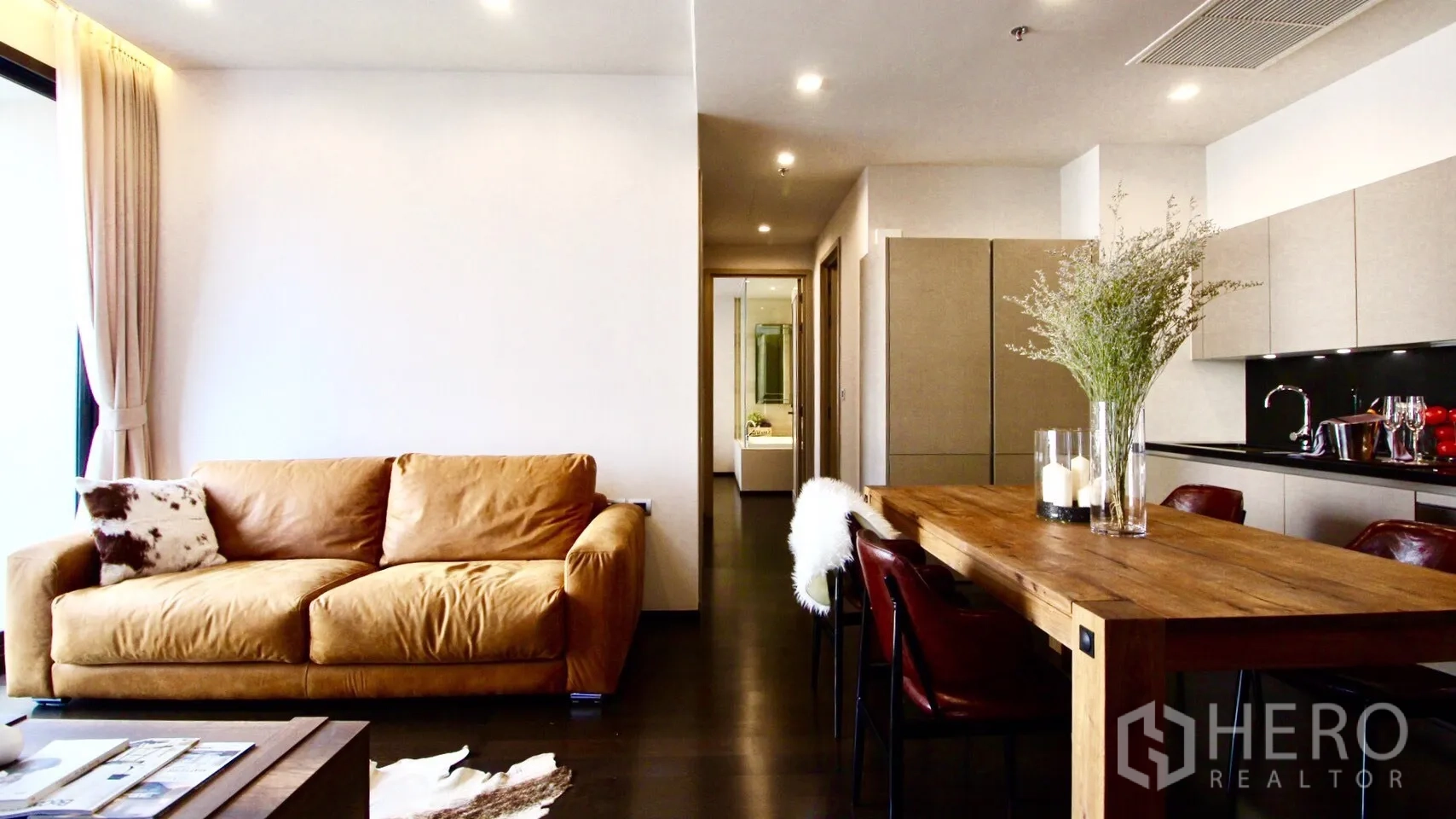 Condominium for rent or sale in Watthana, Bangkok - Open living and dining space with leather sofa, long wooden table, and full kitchen cabinetry.