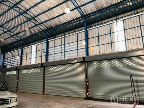 Warehouse for rent in Nakhon Pathom - Row of metal roller shutters beneath tall windows and blue steel trusses inside the warehouse.