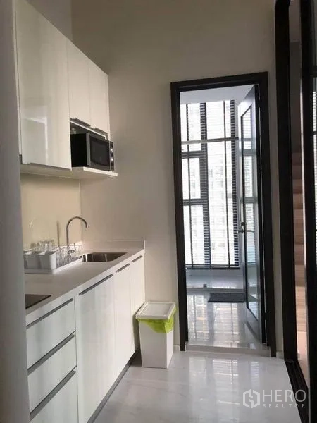 Condominium for sale in Ratchathewi, Bangkok - Compact white kitchenette with built-in cabinets, sink and microwave opening to a glass-walled balcony area.