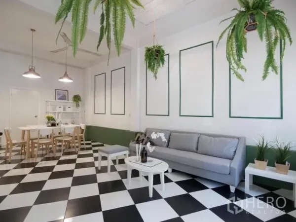 Townhouse for rent in Khlong Toei, Bangkok - Spacious lounge with a long gray sofa, pendant lights, decorative wall panels and checkered tiles leading to the dining corner.