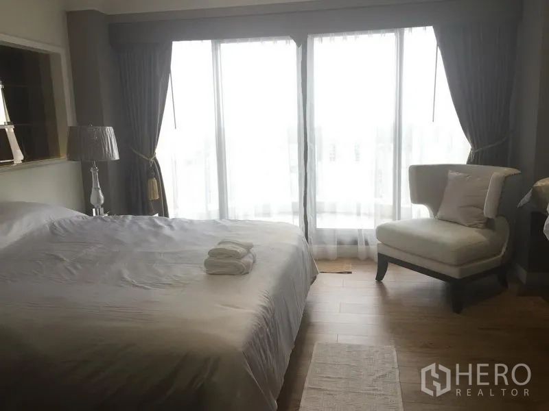Condominium for rent in Bang Rak, Bangkok - Sunlit bedroom with king bed, armchair and floor-to-ceiling windows with curtains.