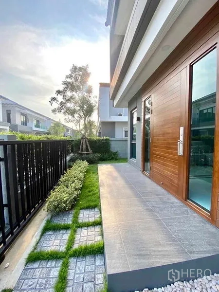 Detached House for rent or sale in Bang Phli, Samut Prakan - Front terrace with tiled patio, wooden entry door, landscaped garden and metal fence.