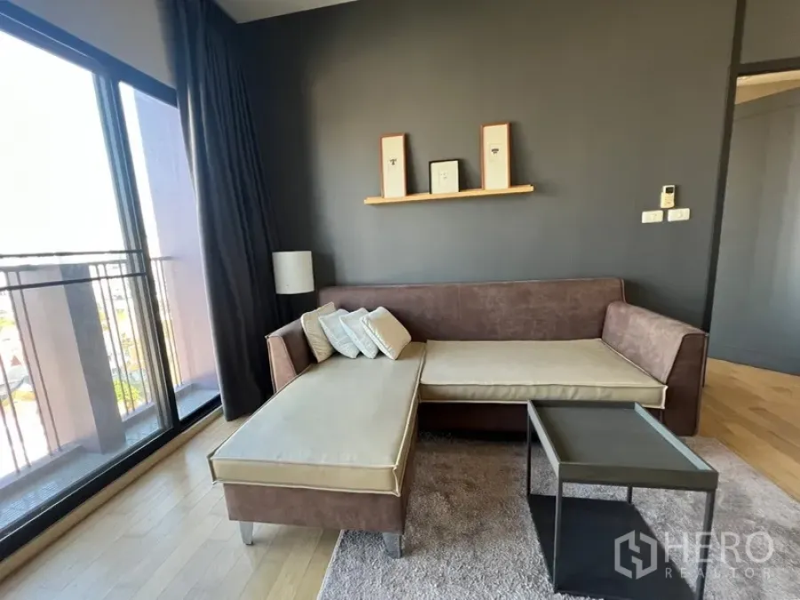 Condominium for rent or sale in Watthana, Bangkok - Bright living room with L-shaped sofa, balcony doors and a dark feature wall at Noble Reveal