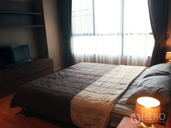 Condominium for rent or sale in Mueang Samut Prakan, Samut Prakan - Bright master bedroom with large window, wood floors, TV console and bedside lamp.