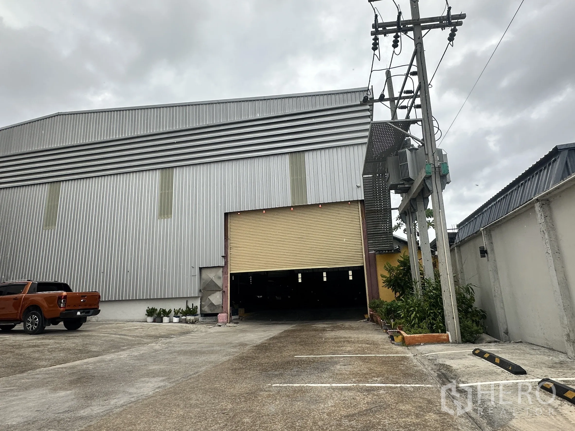 Warehouse for rent in Mueang Samut Prakan, Samut Prakan - Entrance with rolling shutter, parking bays and boundary wall around the site.