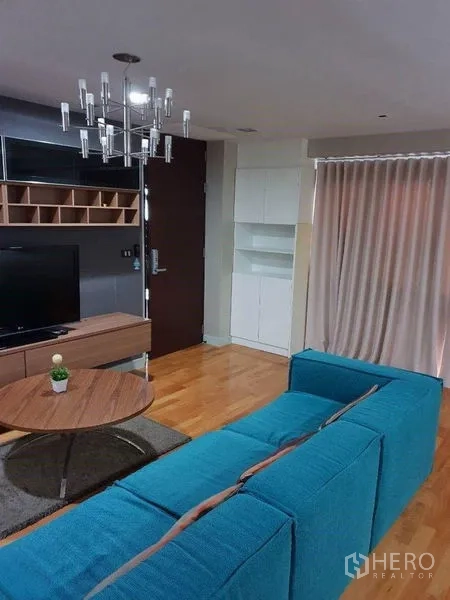 Condominium for sale in Bang Rak, Bangkok - Living area with chandelier, curtains and teal sofa facing TV cabinet.