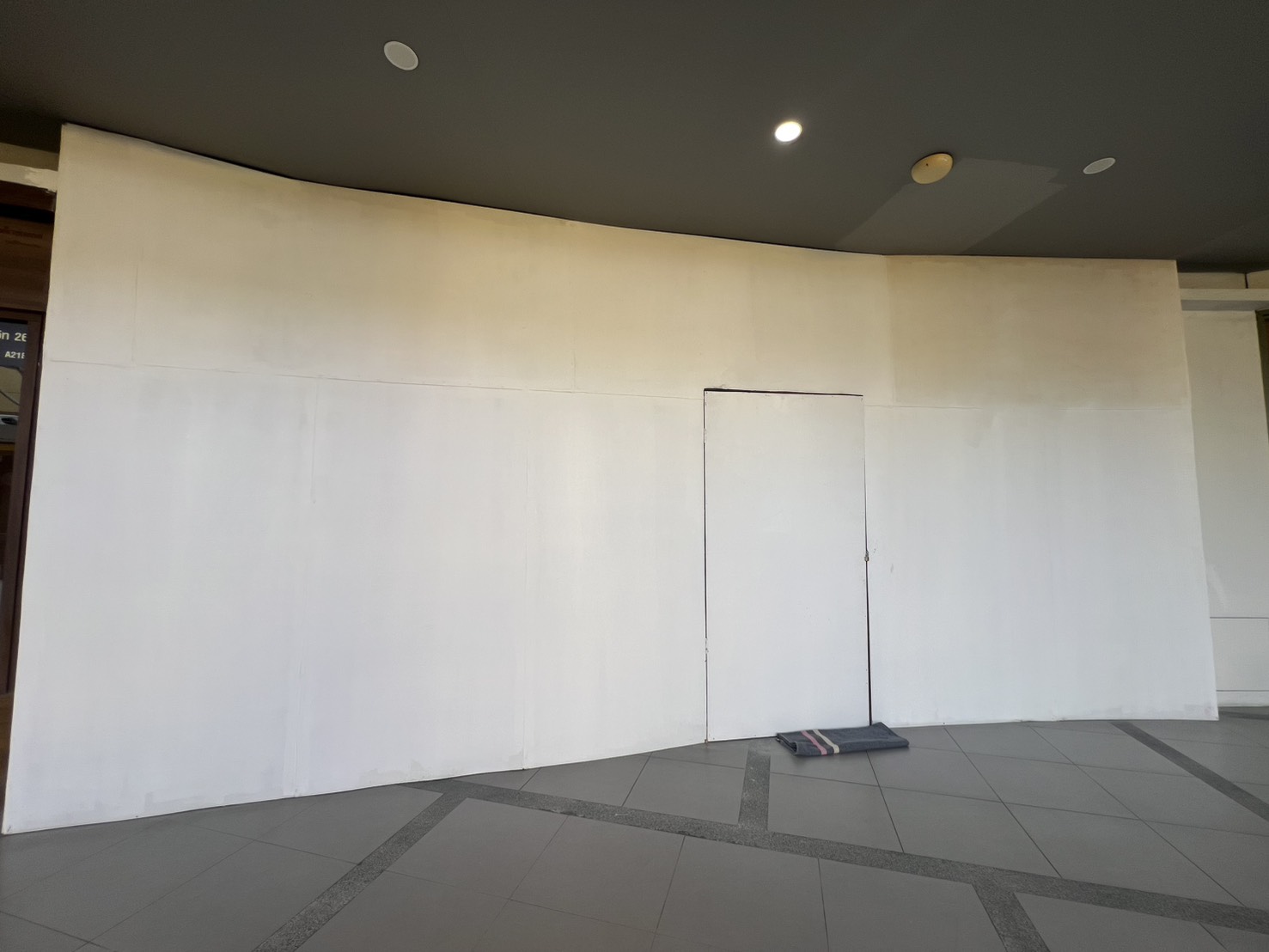 Retail Space for rent in Khlong Toei, Bangkok - Vacant retail unit frontage with a white curved hoarding and small access door beside a tiled indoor walkway with recessed ceiling lights.