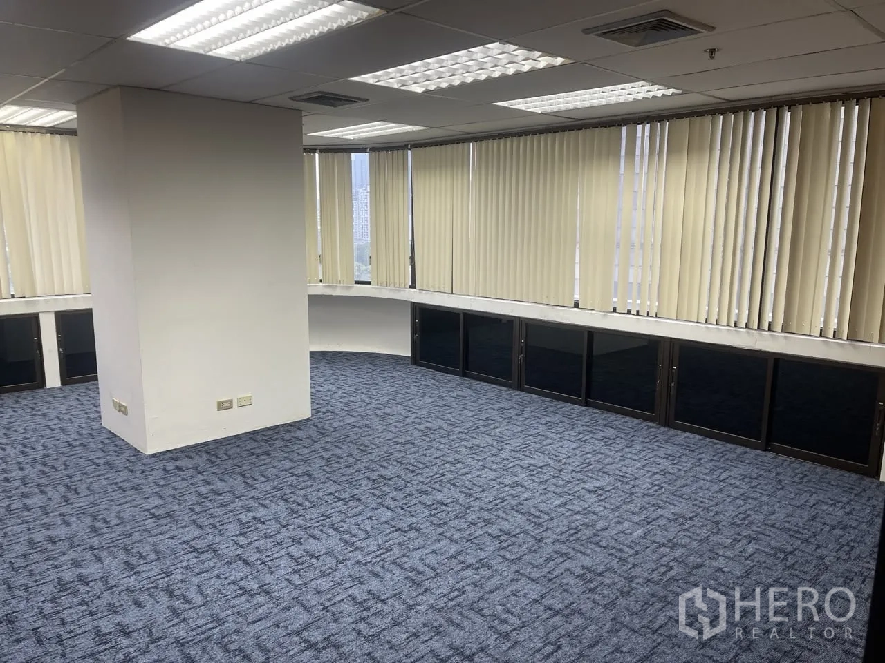Office Space for rent in Khlong Toei, Bangkok - Corner office with blue carpet, central column, drop ceiling lights, and vertical blinds along curved windows.