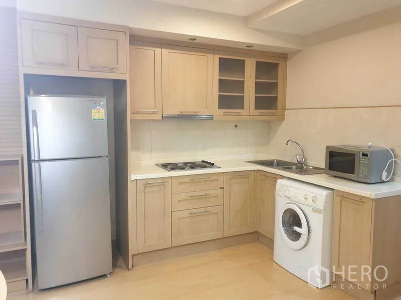 Condominium for rent in Watthana, Bangkok - U-shaped light-wood kitchen featuring cooktop, stainless fridge, microwave, sink, and in-unit washing machine.