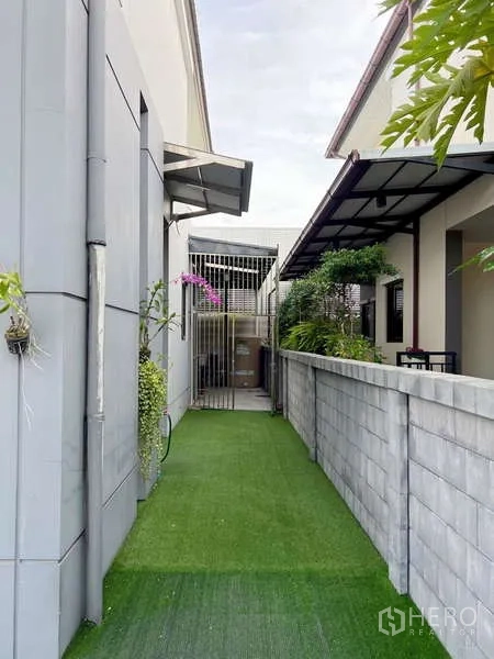 Detached House for sale in Prawet, Bangkok - Narrow side garden walkway with artificial turf, privacy wall, and shaded service area.