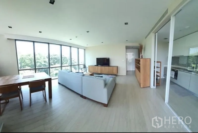Condominium for rent or sale in Watthana, Bangkok - Bright open-plan living and dining room with floor-to-ceiling windows, L-shaped sofa and TV console beside a glass-partitioned kitchen in a Watthana condo.