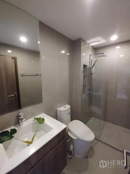 Condominium for rent in Huai Khwang, Bangkok - Modern bathroom with glass-enclosed shower, vanity, and neutral tiled walls.