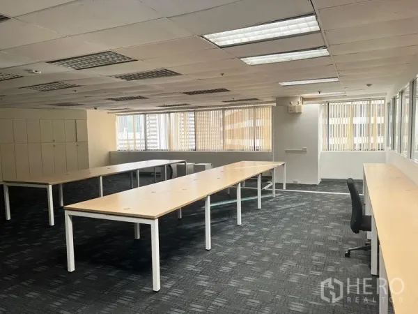 Office Space for rent in Khlong Toei, Bangkok - Open-plan office with long wooden desks, carpet tiles, bright fluorescent ceiling lights, and large windows with vertical blinds.