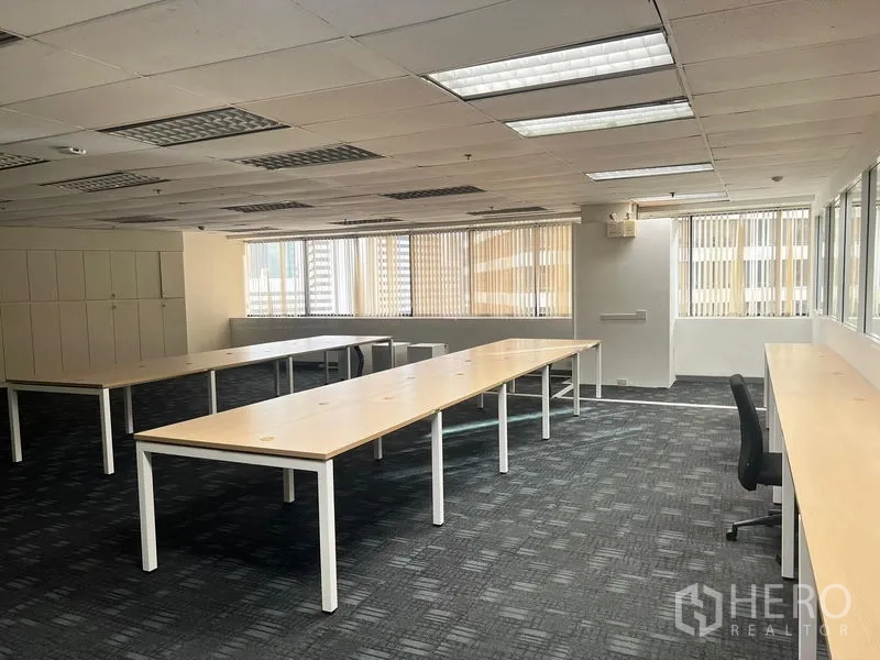 Office Space for rent in Khlong Toei, Bangkok - Open-plan office with long wooden desks, carpet tiles, bright fluorescent ceiling lights, and large windows with vertical blinds.