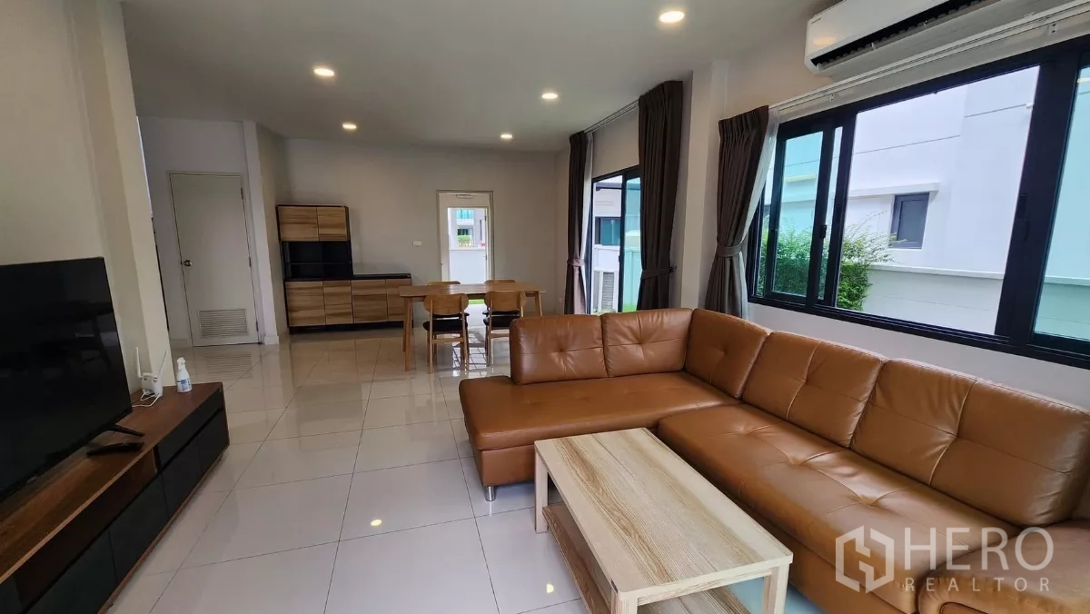 Detached House for rent in Bang Phli, Samut Prakan - Open-plan lounge with sectional sofa, dining area in the background, tiled floors and large windows with curtains.