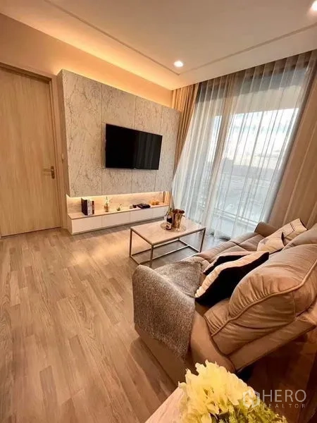 Condominium for sale in Bang Sue, Bangkok - Cozy living area with beige sofa, marble TV feature wall, sheer curtains and balcony access.