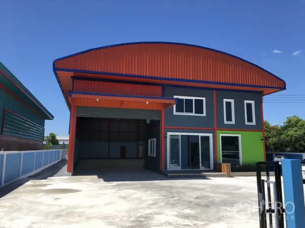 货栈 出租或出售在Sai Noi, Nonthaburi - 现代仓库外观，橙色屋顶，宽幅卷闸门，玻璃办公立面与宽阔混凝土前院。