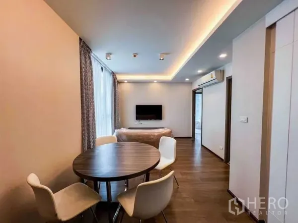 Condominium for sale in Huai Khwang, Bangkok - Open-plan living and dining area with a round table, four white chairs, wall-mounted TV, cove lighting and warm wood floors.