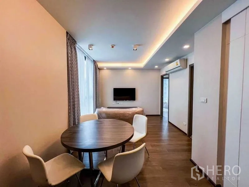 Condominium for sale in Huai Khwang, Bangkok - Open-plan living and dining area with a round table, four white chairs, wall-mounted TV, cove lighting and warm wood floors.