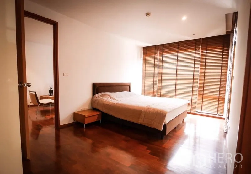 Condominium for rent in Pathum Wan, Bangkok - Spacious bedroom with double bed, bedside table and wall-to-wall windows with wooden blinds over wood floors.