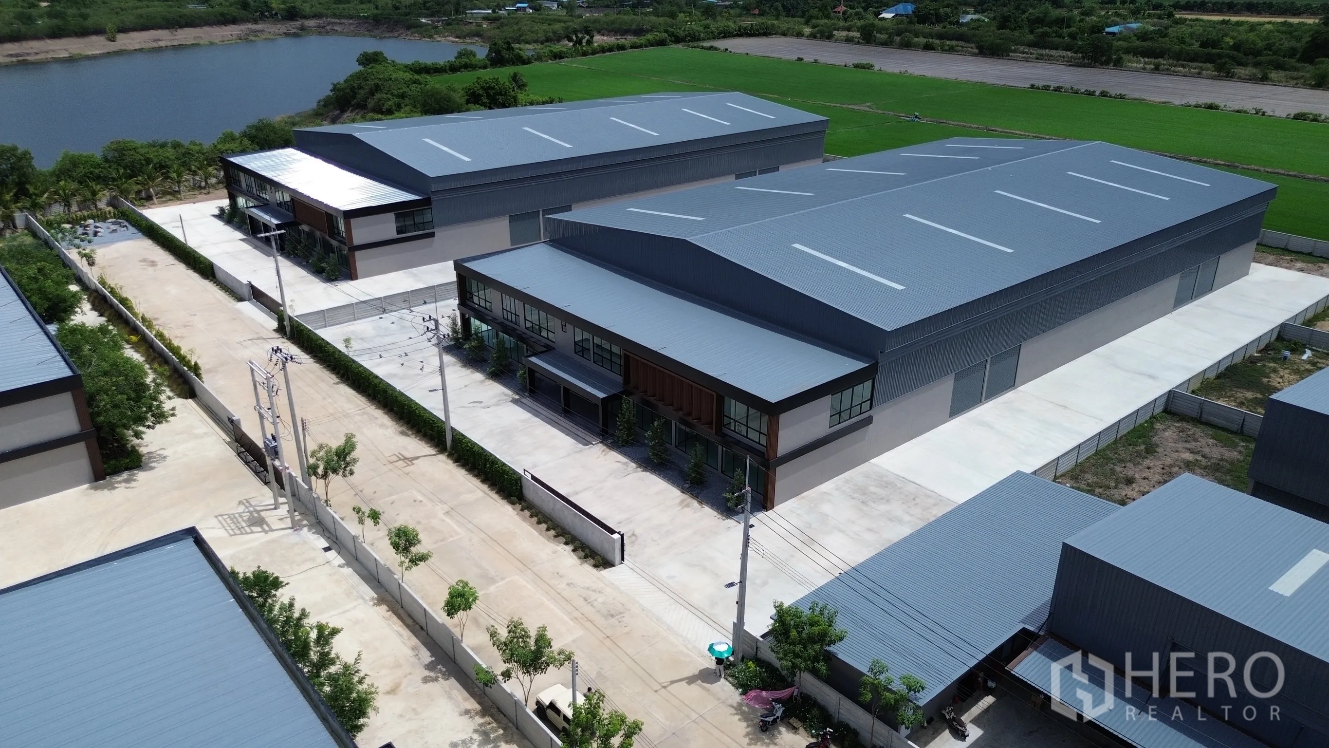 Factory for rent or sale in Bang Len, Nakhon Pathom - Aerial view of multiple warehouse buildings with gray metal roofs, internal roads, perimeter walls, and nearby green fields and water.