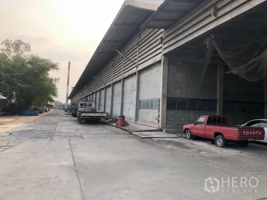 工厂 出租在Bang Bua Thong, Nonthaburi - 厂房外立面沿街延伸，带遮雨装卸区与停靠卡车的宽阔车道