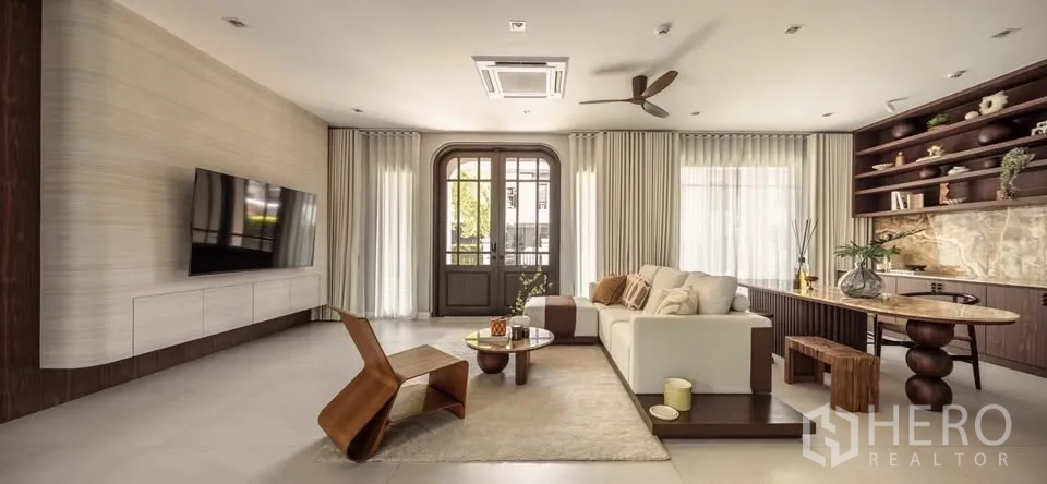 Detached House for rent or sale in Lat Krabang, Bangkok - Bright living room with mounted TV, sectional sofa, built-in shelving, and tall windows.
