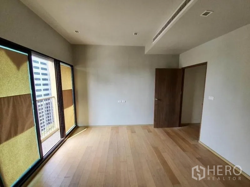 Condominium for sale in Ratchathewi, Bangkok - Bright unfurnished living area with light wood floors and sliding doors to a small balcony overlooking city buildings.