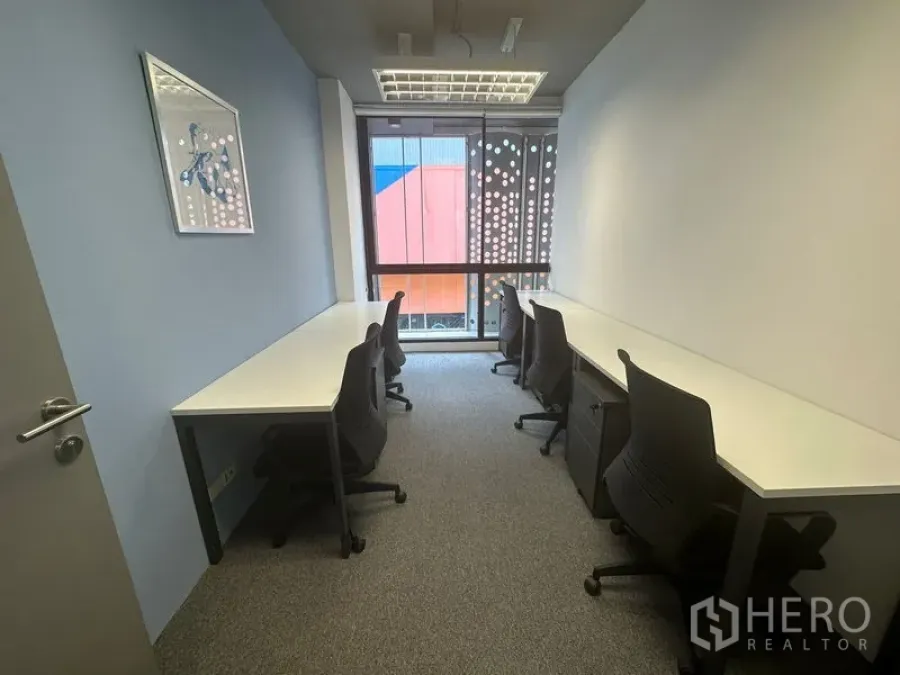 Office Space for rent - Narrow office with long workbenches on both sides, rolling chairs, carpet and floor-to-ceiling window.