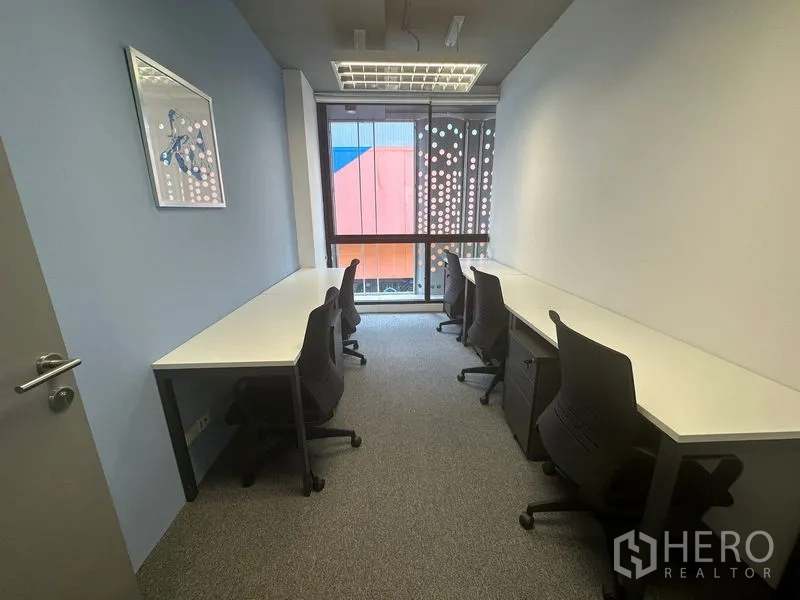 Office Space for rent - Narrow office with long workbenches on both sides, rolling chairs, carpet and floor-to-ceiling window.