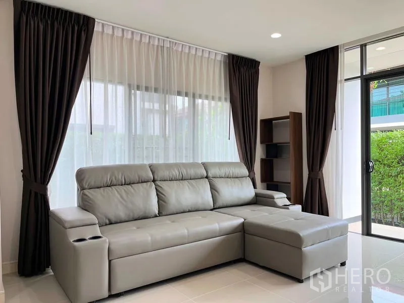 Detached House for rent in Bang Kapi, Bangkok - Living room with L-shaped leather sofa, sheer curtains and sliding door to garden