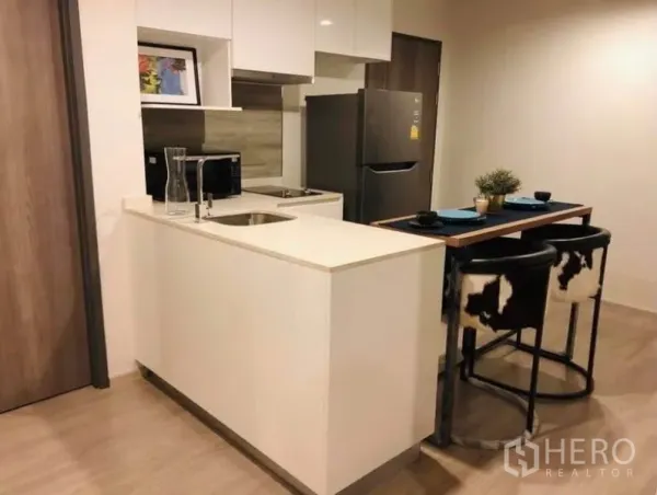 Condominium for rent or sale in Pathum Wan, Bangkok - Modern kitchenette with white peninsula counter, sink, refrigerator and breakfast bar stools.
