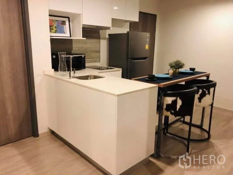 Condominium for rent or sale in Pathum Wan, Bangkok - Modern kitchenette with white peninsula counter, sink, refrigerator and breakfast bar stools.