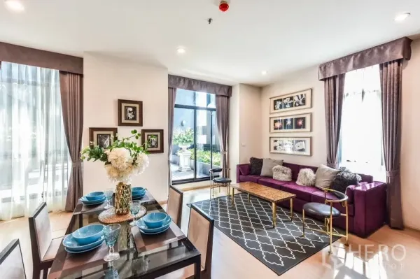 Condominium for rent or sale in Huai Khwang, Bangkok - Bright open-plan dining and living room with a purple sofa, glass dining table, and sliding doors to a greenery-lined terrace.