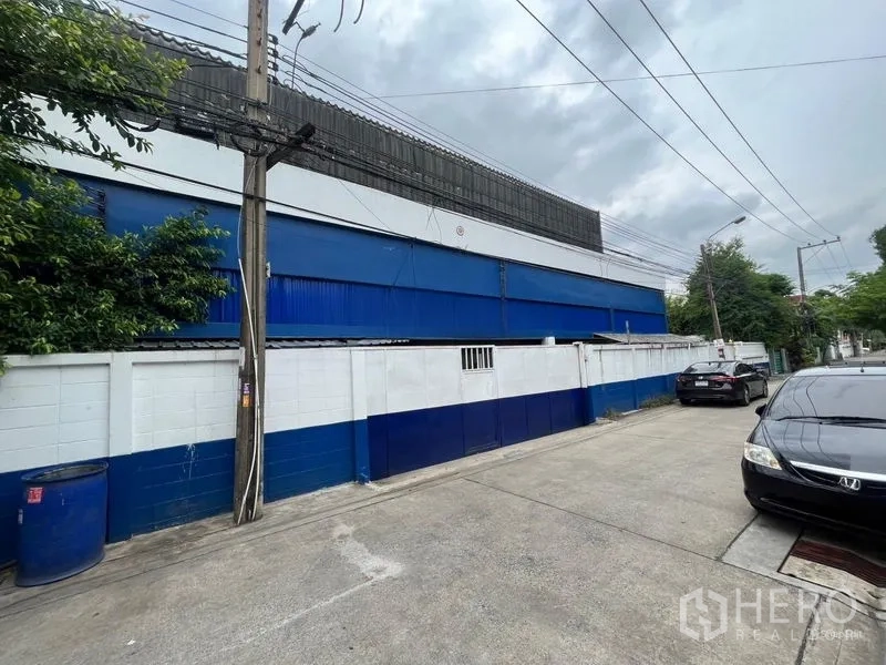 Warehouse for rent in Phra Khanong, Bangkok - Street view of blue-and-white walled warehouse with parked cars and power lines.