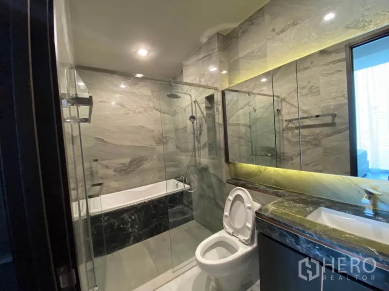 Condominium for rent in Watthana, Bangkok - Luxury bathroom with marble tiles, glass-enclosed shower, bathtub and illuminated vanity.