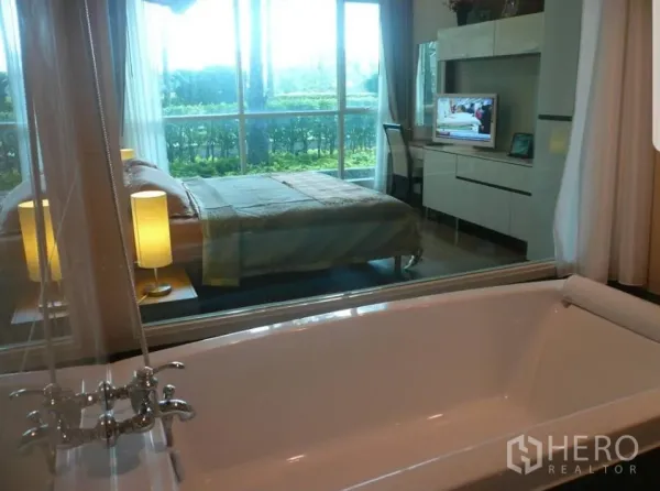 公寓 出租在Pathum Wan, Bangkok - 从浴缸透过玻璃隔断看到卧室，带电视柜与书桌，卧室连接观景阳台与绿篱。