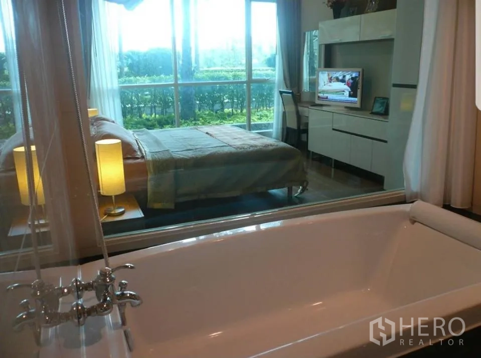 公寓 出租在Pathum Wan, Bangkok - 从浴缸透过玻璃隔断看到卧室，带电视柜与书桌，卧室连接观景阳台与绿篱。
