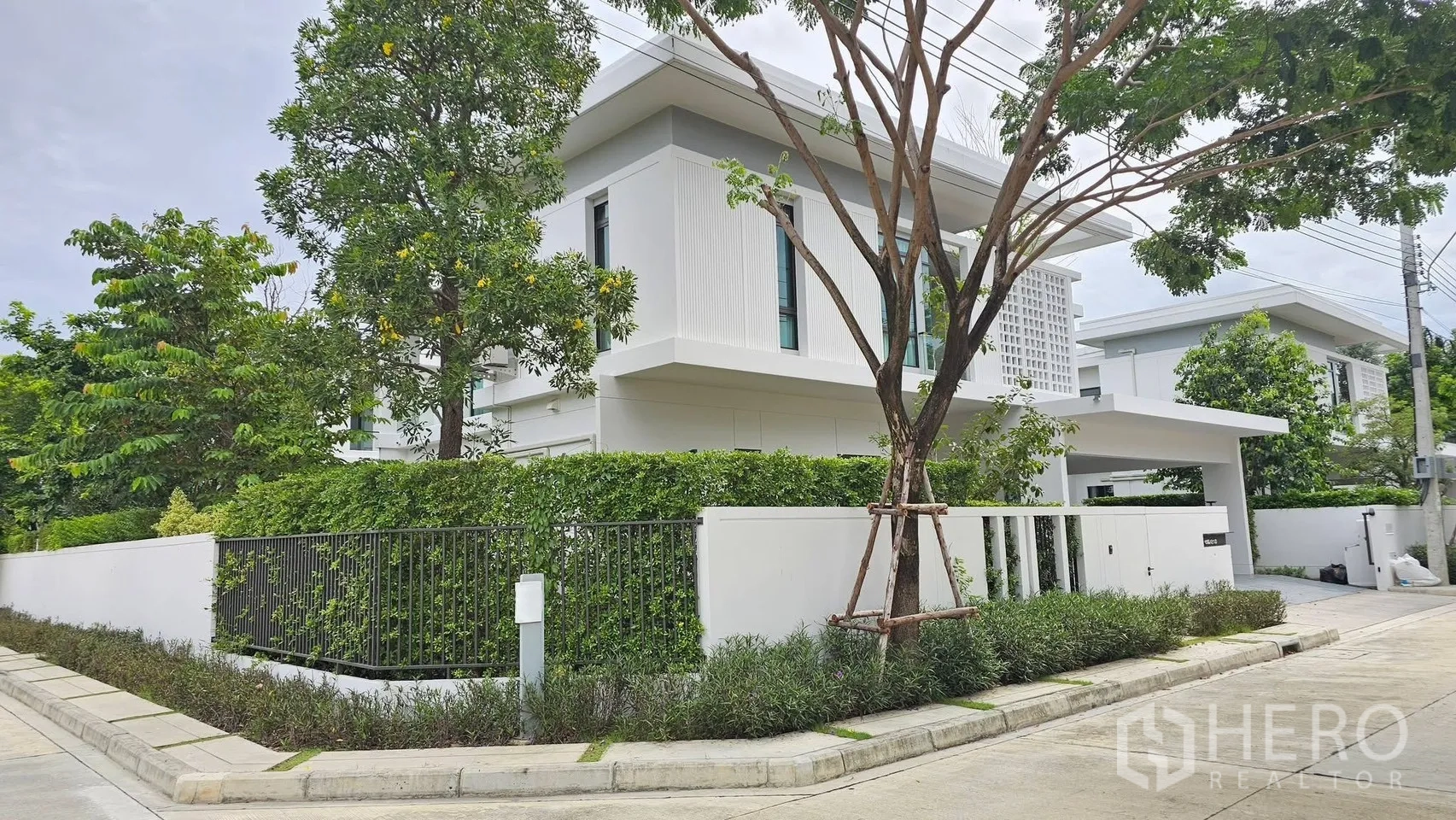 บ้านเดี่ยว for rent in Bang Phli, Samut Prakan - บ้านเดี่ยวสองชั้นสีขาวสมัยใหม่ รายล้อมด้วยต้นไม้และรั้วพุ่มไม้รอบบ้าน.