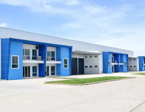 工厂 出租在Bang Sao Thong, Samut Prakan - 现代蓝白色工厂外观，带办公区、卷闸门和宽阔车道的工业园区一角